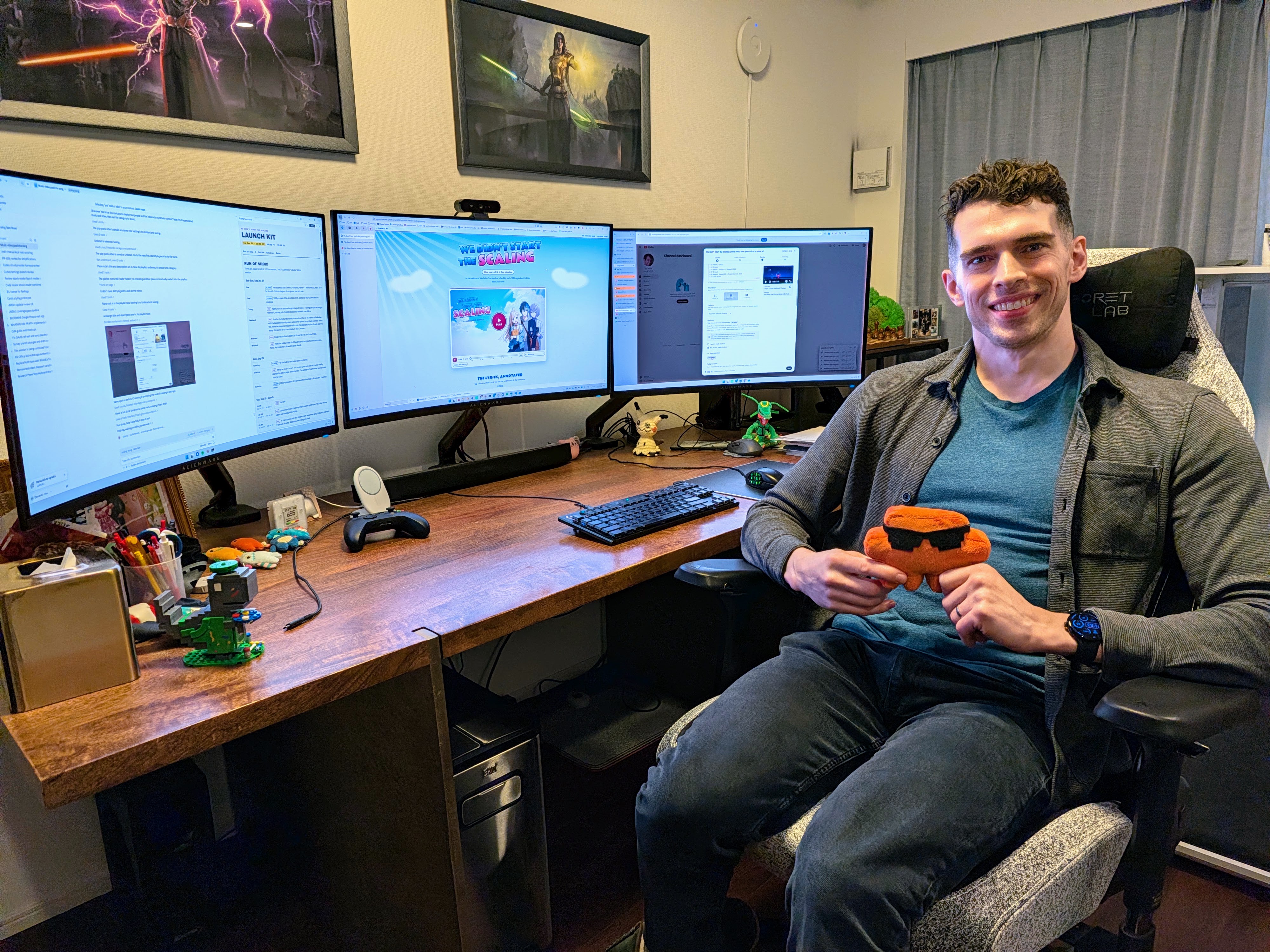 Me at my home office desk holding a Clawd plushie, with three monitors in the background displaying Claude busily prepping the site for publication